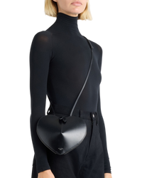 Alaïa Le Coeur leather shoulder bag in black on model, worn crossbody on a thin strap over a black turtleneck