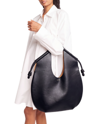 Loewe Paseo knotted leather tote in black worn on the shoulder with a white shirt dress, showing the rounded slouchy shape