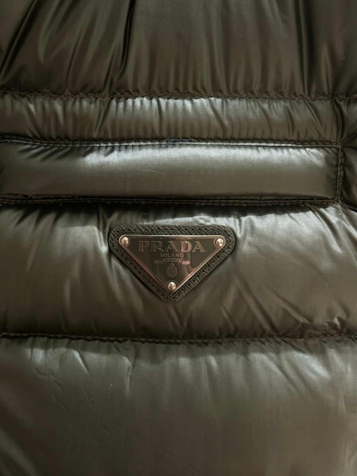 Close-up of the Prada Milano enamelled metal triangle logo on Saffiano leather trim below the zip chest pocket of the green down gilet