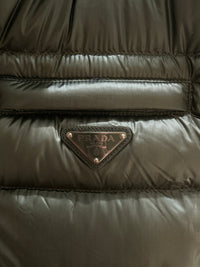 Close-up of the Prada Milano enamelled metal triangle logo on Saffiano leather trim below the zip chest pocket of the green down gilet
